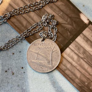 May include: Silver necklace with a coin pendant. The coin is inscribed with "REPUBBLICA ITALIANA" and the year "1953". The chain is a simple, linked design. The pendant is circular with a detailed image.