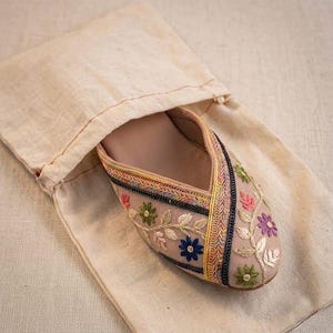 May include: A beige embroidered slipper with floral designs in pink, green, blue, and purple, inside a matching beige drawstring bag. The slipper has a decorative trim in multiple colors.