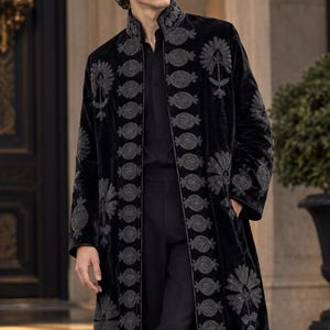 May include: A long black velvet coat featuring a high collar and intricate grey floral and geometric embroidery along the front opening and hem. The coat is worn over a black button-down shirt and black trousers, creating a dark, layered ensemble.