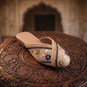 May include: A beige slipper with floral embroidery. The slipper features a tan sole and a sheer overlay adorned with colorful embroidered flowers. The design includes a cross-strap detail with decorative stitching. The slipper is displayed on a carved wooden surface.