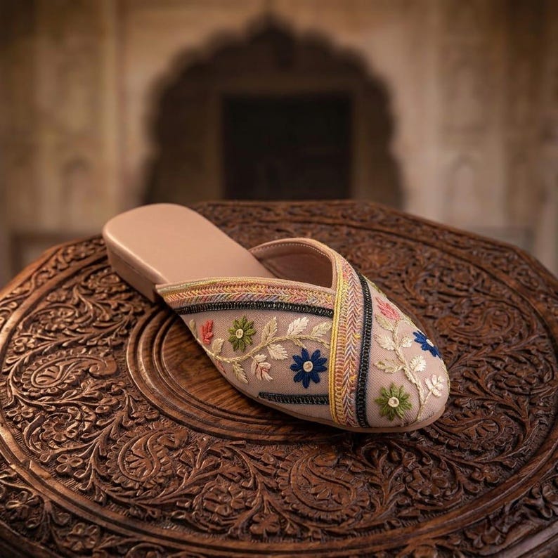 May include: A beige slipper with floral embroidery. The slipper features a tan sole and a sheer overlay adorned with colorful embroidered flowers. The design includes a cross-strap detail with decorative stitching. The slipper is displayed on a carved wooden surface.