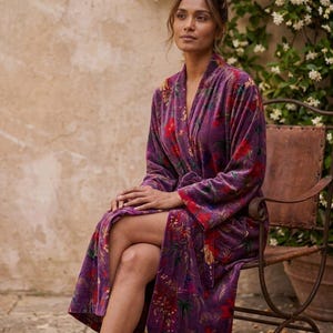 May include: A long, purple velvet robe with a floral pattern in red and green. The robe is open, revealing the legs of the person wearing it. The person is seated on a brown leather chair.