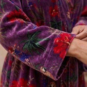May include: Close-up of a purple velvet robe with a vibrant floral pattern. The design features large red hibiscus flowers, green palm leaves, and blue accents. The robe's cuff showcases a parrot illustration.