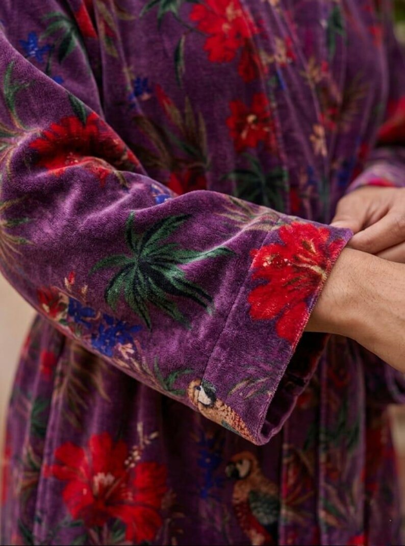 May include: Close-up of a purple velvet robe with a vibrant floral pattern. The design features large red hibiscus flowers, green palm leaves, and blue accents. The robe's cuff showcases a parrot illustration.