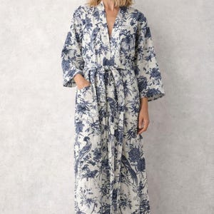 May include: A long, white robe featuring a blue floral and bird design. The robe has a tie waist, pockets, and three-quarter length sleeves. The fabric looks lightweight and comfortable, suitable for lounging.