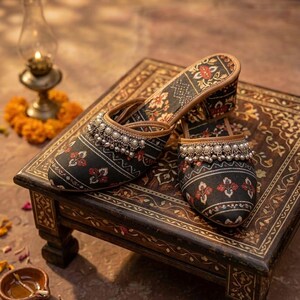 Puede incluir: Un par de zapatos sin cordones con estampado negro y rojo con adornos de cuentas plateadas. Los zapatos se exhiben sobre una peque&ntilde;a mesa de madera ornamentada con un dise&ntilde;o floral. El fondo incluye una l&aacute;mpara de aceite encendida y p&eacute;talos de flores.