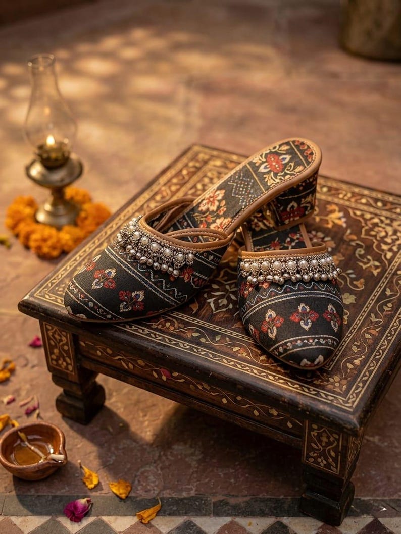 Puede incluir: Un par de zapatos sin cordones con estampado negro y rojo con adornos de cuentas plateadas. Los zapatos se exhiben sobre una peque&ntilde;a mesa de madera ornamentada con un dise&ntilde;o floral. El fondo incluye una l&aacute;mpara de aceite encendida y p&eacute;talos de flores.