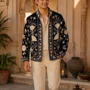 May include: A black jacket featuring intricate white floral embroidery along the front panels and collar, worn over a light beige button-down shirt and matching light beige trousers, paired with brown leather sandals.