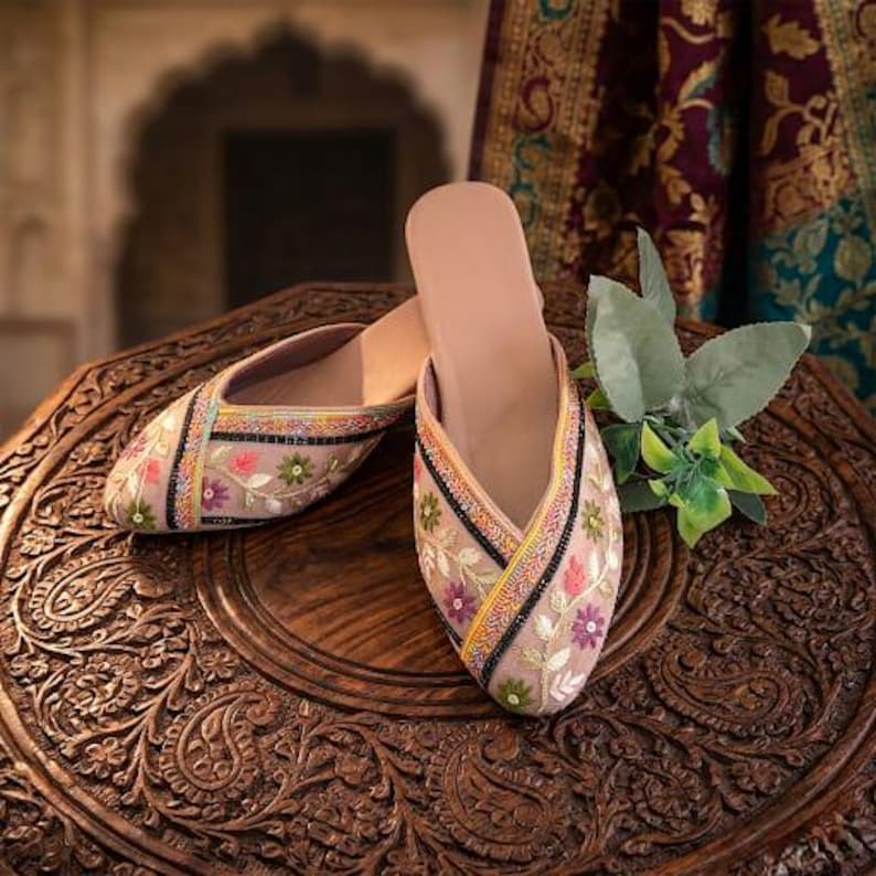 May include: A pair of beige embroidered slip-on shoes with floral patterns in pink, green, and purple. The shoes have a black border and are displayed on a carved wooden table. The background features a draped fabric.