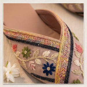 May include: Close-up of a blush-colored flat shoe with intricate floral embroidery. The shoe features a delicate mesh overlay adorned with colorful stitched flowers and leaves. The edges are trimmed with a detailed, multi-colored border. A white flower is in the lower left corner.