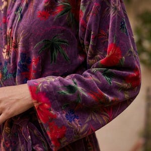 May include: A plush, purple robe with a vibrant floral pattern. The design features red, green, and blue flowers and foliage against a deep purple background. The robe's sleeves and edges are trimmed with a contrasting floral border.