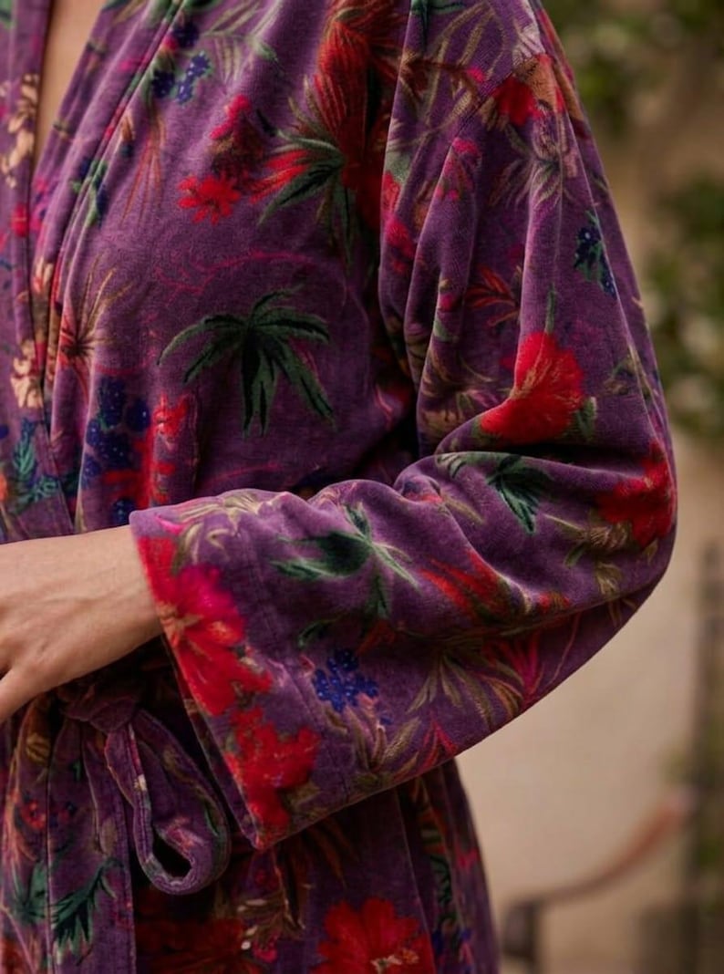 May include: A plush, purple robe with a vibrant floral pattern. The design features red, green, and blue flowers and foliage against a deep purple background. The robe's sleeves and edges are trimmed with a contrasting floral border.