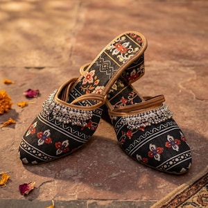 Puede incluir: Un par de zapatos negros sin cordones con un estampado floral en rojo, blanco y dorado. Los zapatos tienen un adorno de cuentas plateadas en la parte superior y un ribete de cuero marr&oacute;n. Los zapatos se exhiben sobre una superficie de baldosas marrones.