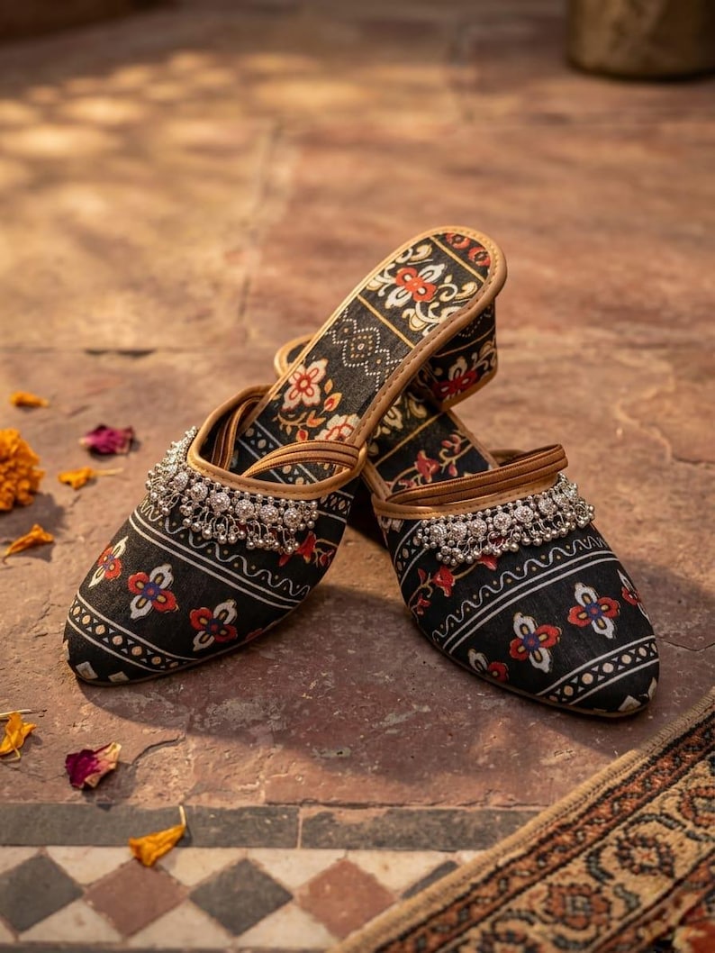 Puede incluir: Un par de zapatos negros sin cordones con un estampado floral en rojo, blanco y dorado. Los zapatos tienen un adorno de cuentas plateadas en la parte superior y un ribete de cuero marr&oacute;n. Los zapatos se exhiben sobre una superficie de baldosas marrones.