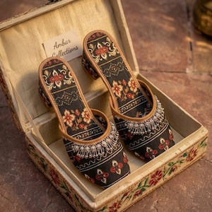Puede incluir: Un par de zapatos con estampado floral negro y rojo con tacones peque&ntilde;os, exhibidos dentro de una caja decorativa. Los zapatos tienen un ribete de cuentas y est&aacute;n etiquetados como "Ambar Collections". La caja es beige con dise&ntilde;os florales.