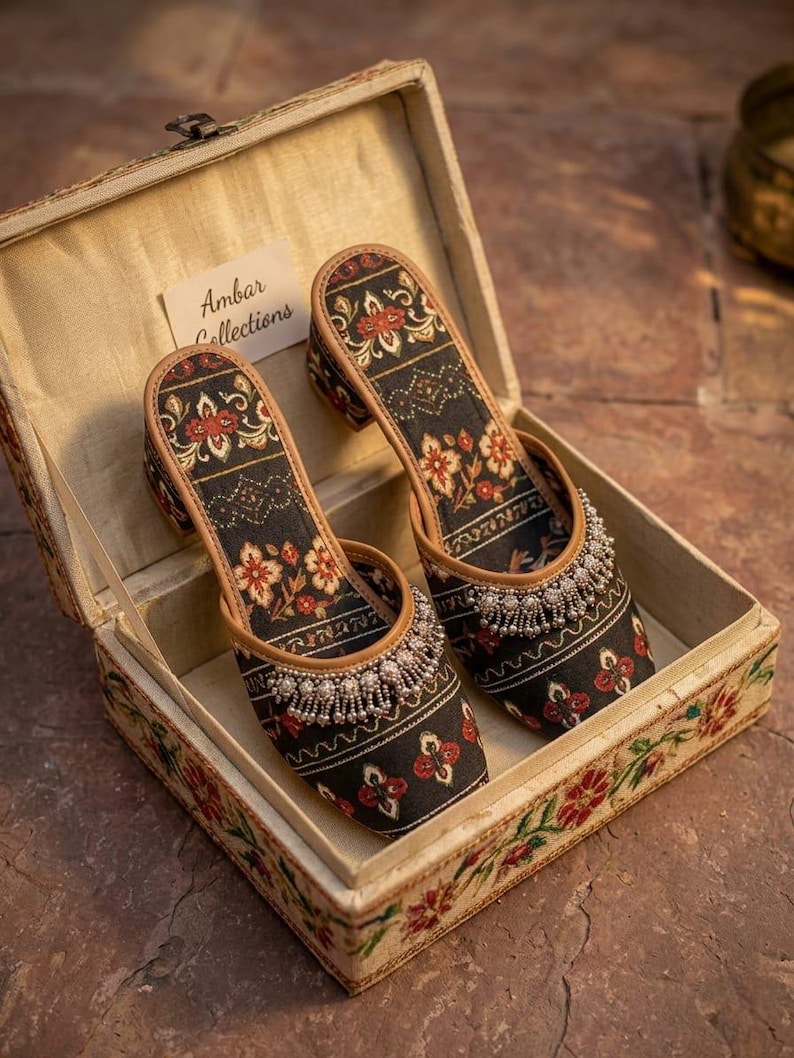 Puede incluir: Un par de zapatos con estampado floral negro y rojo con tacones peque&ntilde;os, exhibidos dentro de una caja decorativa. Los zapatos tienen un ribete de cuentas y est&aacute;n etiquetados como "Ambar Collections". La caja es beige con dise&ntilde;os florales.