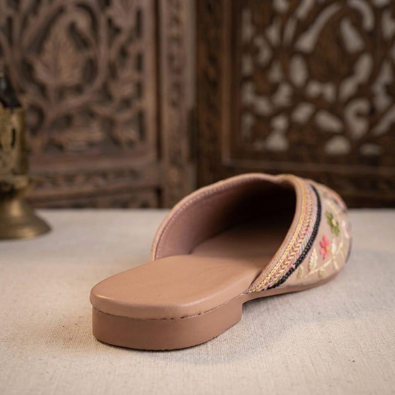 May include: A beige mule slipper with a flat sole. The slipper features embroidered floral designs in pink, green, and yellow. The trim includes gold and black accents. The interior is a light pink color.