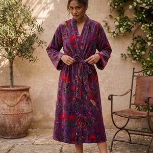 May include: A long, purple floral robe with a tie waist. The robe features a vibrant pattern of red and green flowers on a dark purple background. The robe is made of a soft, velvety material and has wide sleeves.
