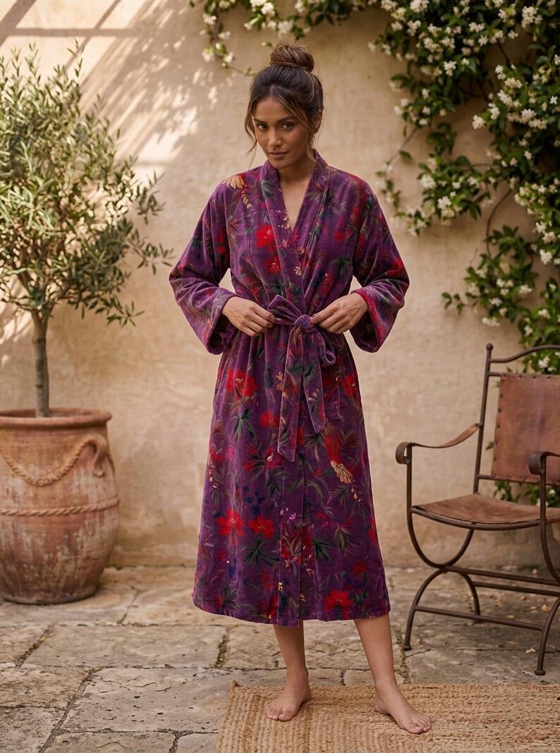 May include: A long, purple floral robe with a tie waist. The robe features a vibrant pattern of red and green flowers on a dark purple background. The robe is made of a soft, velvety material and has wide sleeves.