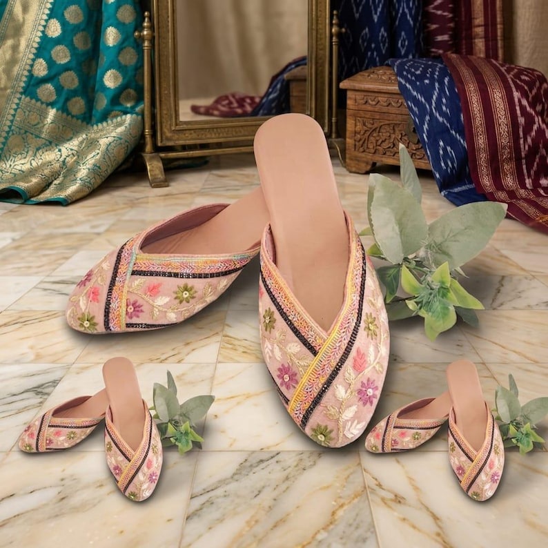May include: A pair of blush-colored embroidered slippers with floral and geometric patterns. The slippers have a pointed toe and a flat sole. The embroidery includes pink, green, and yellow flowers with black and gold accents. Additional pairs are also shown.
