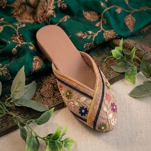 May include: A beige slip-on shoe with floral embroidery in pink, blue, and green. The shoe features a decorative trim with black, yellow, and red accents. The shoe is set against a backdrop of green fabric with gold floral patterns.