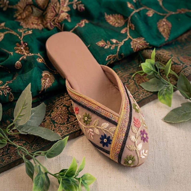 May include: A beige slip-on shoe with floral embroidery in pink, blue, and green. The shoe features a decorative trim with black, yellow, and red accents. The shoe is set against a backdrop of green fabric with gold floral patterns.