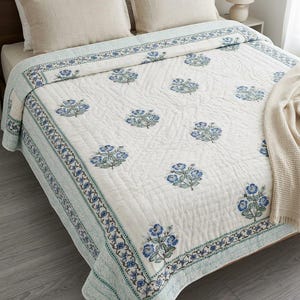 Op de afbeelding: Een wit gewatteerd sprei met een bloemmotief in blauw- en groentinten. Het sprei heeft een decoratieve rand en ligt op een bed met beige kussens. Een beige sprei is over het bed gedrapeerd.