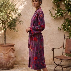 May include: A long, purple robe with a floral pattern featuring red and green accents. The robe is tied at the waist and has long sleeves. The image is taken outdoors, with a stone patio and a potted tree in the background.