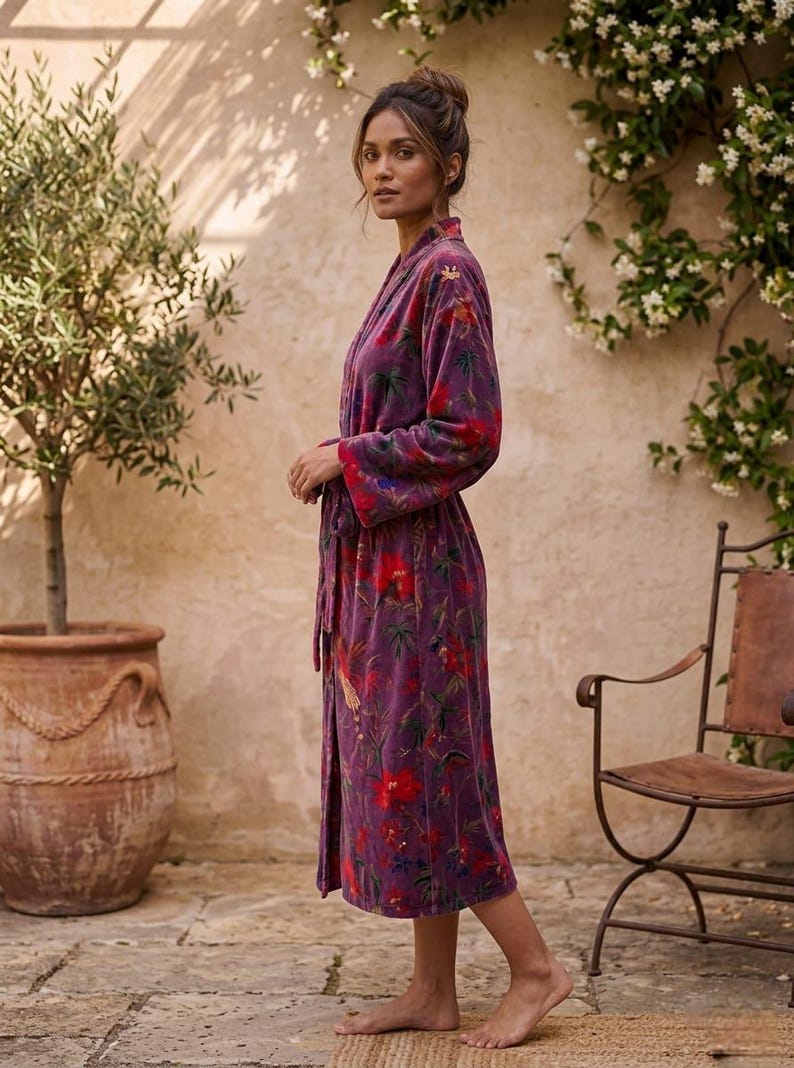 May include: A long, purple robe with a floral pattern featuring red and green accents. The robe is tied at the waist and has long sleeves. The image is taken outdoors, with a stone patio and a potted tree in the background.