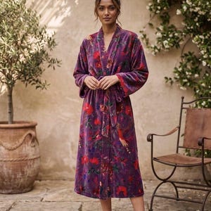 Pode incluir: Robe longa de veludo roxo com um padrão floral com detalhes em vermelho e verde. O roupão tem uma cintura com laço e mangas largas. A modelo está em um jardim com piso de pedra e uma planta em vaso.