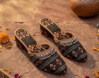 Op de afbeelding: Een paar zwarte en rode muiltjes met bloemenpatroon en een kleine blokhak. De schoenen hebben een zilveren kralenversiering aan de bovenkant en een bruinleren rand. De schoenen worden op een stenen oppervlak gepresenteerd.