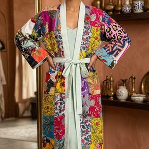May include: A long, patchwork-style robe with a light green tie belt. The robe features a variety of colorful floral and patterned fabrics. The robe is worn over a light green dress. The robe's trim is a light green and white stripe.