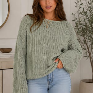 May include: A light green knit sweater with long sleeves and a boat neck. The sweater is paired with light blue jeans. The woman is wearing a gold necklace and hoop earrings. A round mirror and a small plant are in the background.