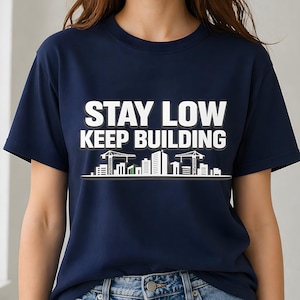 May include: Navy blue t-shirt with the text "STAY LOW KEEP BUILDING" in white block letters. Below the text is a white line drawing of a city skyline with construction cranes and buildings. The shirt is a crew neck.