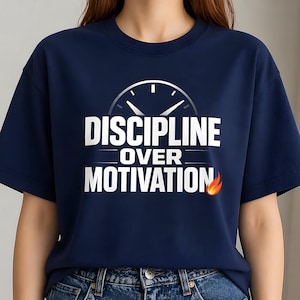 Può includere: T-shirt blu navy con la scritta bianca "DISCIPLINE OVER MOTIVATION", una grafica a orologio e una fiamma. La maglietta è a girocollo.