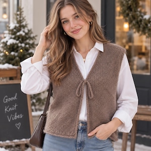 May include: A brown knitted vest featuring a V-neckline and a front tie closure, worn over a crisp white long-sleeved button-down shirt and light blue denim jeans. The vest has a simple, textured knit pattern and a straight hemline at the waist.