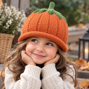 May include: A knitted orange pumpkin hat with a green stem and leaf-shaped detailing at the top. The hat features a ribbed texture throughout the main body and a rolled brim at the bottom edge, crafted from yarn in a vibrant pumpkin orange color.
