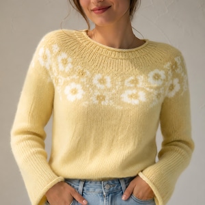 Könnte beinhalten: Ein blassgelber Langarmpullover mit einem weißen Blumenmuster um den Halsausschnitt und die Schultern. Das Strickkleidungsstück hat einen gerippten Rundhalsausschnitt und gerippte Bündchen, kombiniert mit einer hellblauen Jeans mit sichtbarem Metallknopf.