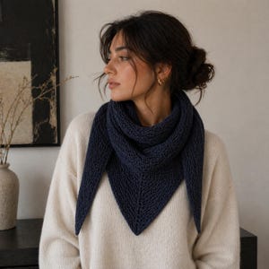 May include: A dark navy blue knitted scarf, with a triangular shape and pointed ends, is worn around the neck. The person is wearing a cream-coloured sweater. The scarf is made of a textured knit fabric.