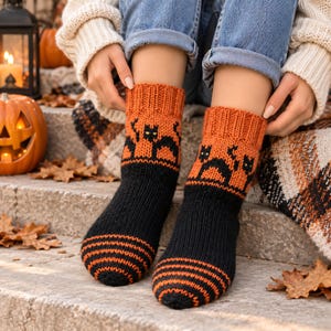 Puede incluir: Un par de calcetines de punto con un diseño en negro y naranja. La sección superior tiene un puño acanalado con un patrón de gatos negros sobre fondo naranja. La sección inferior es de color negro sólido con rayas horizontales naranjas.