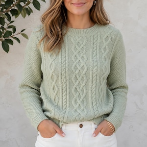 May include: A sage green long-sleeve sweater featuring a prominent vertical cable knit pattern on the front panel, complemented by textured side panels, a ribbed crew neckline, and ribbed cuffs, paired with white pants.