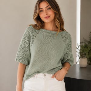 Peut inclure: Un pull en tricot vert clair à manches courtes et col bateau. Les manches présentent un motif ajouré détaillé. Le modèle porte un pantalon blanc et un bracelet doré. Le pull est de couleur vert doux et sourd.