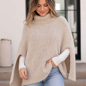 Op de afbeelding: Een beige gebreide poncho met een hoge col en brede armopeningen. Het kledingstuk is gemaakt van een getextureerd, grof garen met een doorlopend ribbelpatroon, wat zorgt voor een losse, gedrapeerde pasvorm over een witte basislaag met lange mouwen.