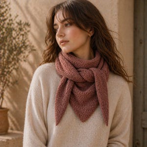 May include: A dusty rose-colored knitted scarf, draped around the neck. The scarf has a textured, open weave design and triangular ends. The person is wearing a cream-colored sweater. The background is a neutral beige.