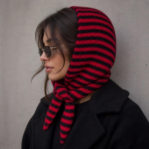 May include: A red and black striped knitted hood scarf. The hood is up, and the scarf is tied under the chin. The person is wearing sunglasses and a black coat. The scarf is made of a soft, warm material.