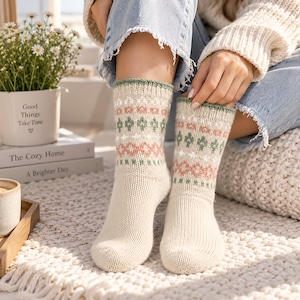 Könnte beinhalten: Ein Paar cremefarbene Strickstrümpfe mit einem sich wiederholenden geometrischen Muster in gedecktem Salbeigrün und Altrosa. Die Socken haben ein geripptes Bündchen am oberen Rand mit einem dünnen salbeigrünen Saum und ein detailliertes Strickmuster.
