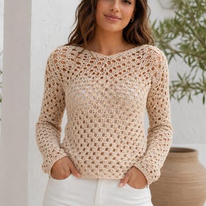 May include: A beige, open-weave crochet sweater with long sleeves and a scoop neck. The sweater has a delicate, airy design, and is paired with white pants. The overall style is casual and relaxed, suitable for warm weather.