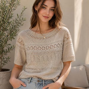 May include: A beige short-sleeved knit sweater featuring a round neckline and a combination of solid stockinette stitch panels and horizontal bands of openwork diamond and geometric patterns throughout the front of the garment.