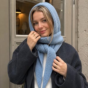 Könnte beinhalten: Ein hellblauer, gestrickter Kapuzenschal mit einer texturierten kraus rechts Strickstruktur. Das Accessoire besteht aus einer Kapuze, die in einen langen Schal übergeht und so um den Hals gewickelt wird, dass zwei spitze Enden nach vorne hängen.