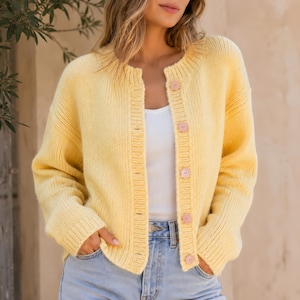 Puede incluir: Un cárdigan de punto amarillo pálido con cuello redondo acanalado, mangas largas con puños acanalados y cierre frontal con cinco botones circulares de color claro. La prenda se lleva sobre una camiseta de tirantes blanca y vaqueros azul claro.